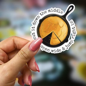 Puede incluir: Una pegatina con un dibujo de un panqueque en una sartén con el texto "cut it down the middle, open wide & jump right in". El panqueque es de color marrón dorado con una porción cortada. La sartén es negra.
