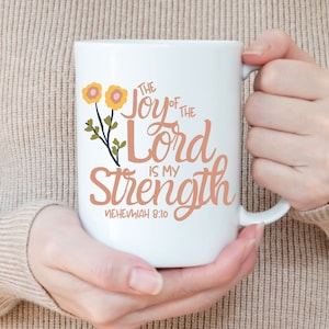 May include: White ceramic mug with a peach-colored floral design and the text "The Joy of the Lord is My Strength" with the scripture reference "Nehemiah 8:10".