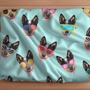 Pode incluir: Um tecido azul e branco com um padrão repetido de um cão usando diferentes óculos de sol. O cão é um blue heeler com o peito branco e marcas brancas no rosto. Os óculos de sol são de várias cores e estilos, incluindo em forma de coração, redondos e retangulares.