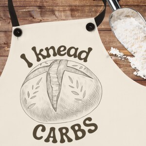 Pode incluir: Um avental branco com um desenho marrom de um pão e o texto "I knead carbs".