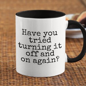May include: White ceramic coffee mug with a black rim and handle. The mug has the text "Have you tried turning it off and on again?" printed in black.