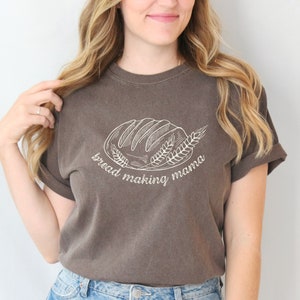 Op de afbeelding: Een bruin t-shirt met een witte afbeelding van een brood en tarwehalmen. De tekst "bread making mama" staat onder de afbeelding.