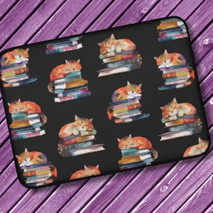 Puede incluir: Una funda para portátil negra con un patrón repetido de gatos naranja y blanco durmiendo sobre pilas de libros coloridos.