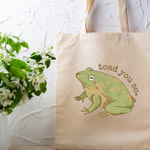 May include: A beige canvas tote bag featuring a green frog illustration and the text "toad you so."