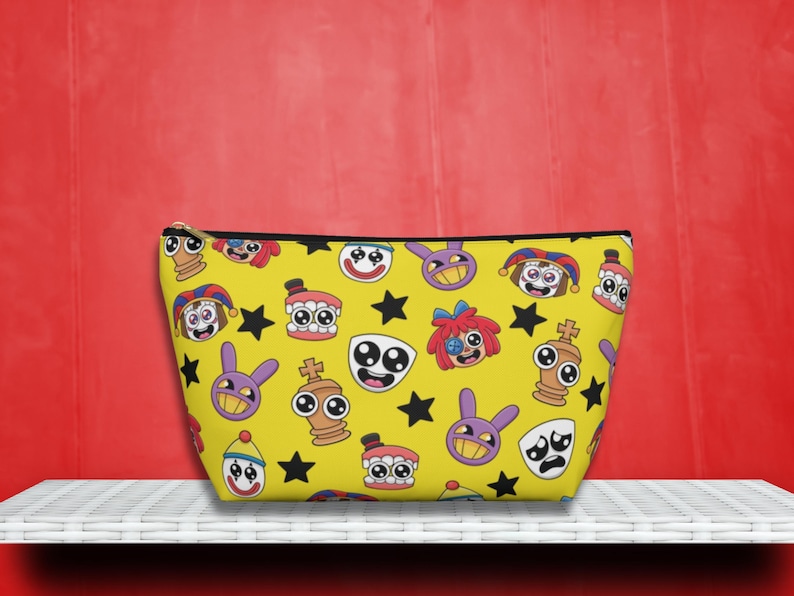 May include: Yellow cosmetic bag with a black zipper and a repeating pattern of cartoon faces, stars, and other whimsical characters. The bag is sitting on a white shelf against a red background.