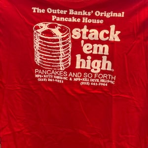 Puede incluir: Camiseta roja con texto blanco que dice "The Outer Banks' Original Pancake House stack 'em high Pancakes and so forth MP4 Kitty Hawk NC & MP9 Kill Devil Hills NC (252) 261-8221 (252) 441-7064". La camiseta presenta un gráfico de una pila de panqueques.