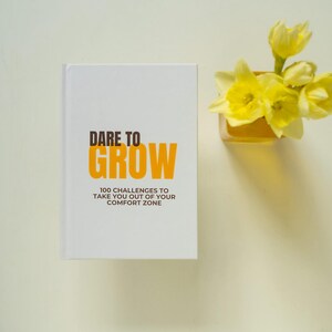 May include: A white book with the title "Dare to Grow" in brown and orange text. The book is on a white surface with a vase of yellow daffodils to the right. The book's subtitle is "100 Challenges to Take You Out of Your Comfort Zone."