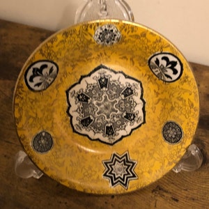 Op de afbeelding: Een geel keramiek bord met een gouden rand en zwarte en witte bloemmotieven. Het bord heeft een centraal ontwerp van een zwarte en witte sneeuwvlok op een witte achtergrond.