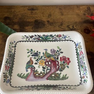 Könnte beinhalten: Rechteckige Servierplatte aus Keramik mit weißem Hintergrund und farbenfrohem Blumen- und Vogeldesign. Die Platte hat einen detaillierten Rand mit einem seilartigen Rand. Das zentrale Design zeigt einen bunten Vogel, der auf einem Ast mit Blumen sitzt.