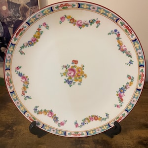 May include: A white porcelain plate with a floral design. The plate has a central floral arrangement and smaller floral clusters around the rim. The rim has a decorative border with red, blue, and yellow accents. The plate is displayed on a black stand.