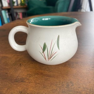 Op de afbeelding: Crèmekleurige keramische creamer met een teal interieur en een kleine tuit. De creamer heeft een afgeronde vorm met een handvat en is versierd met een eenvoudig bloemmotief in groen en rood.