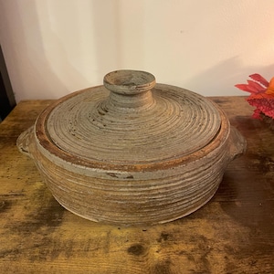 May include: A round, lidded stoneware pot with a textured, ridged design. The pot is a muted grey-brown colour with a small handle on the lid and two side handles. The pot sits on a wooden surface.