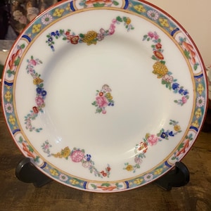 May include: White ceramic plate with a red rim and floral design. The plate features a central floral arrangement and a decorative border with flowers and geometric patterns in various colours, including pink, yellow, and blue.