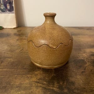 Puede incluir: Un pequeño tarro de cerámica redondo con cuello estrecho y diseño ondulado en el centro. El tarro es de color marrón moteado y se asienta sobre una superficie de madera. El tarro es probablemente hecho a mano y adecuado para guardar objetos pequeños.