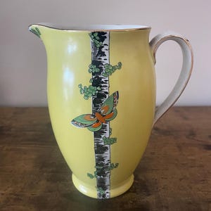 Puede incluir: Jarra de cerámica amarilla con asa blanca y ribete dorado. La jarra presenta un diseño decorativo de un tronco de árbol con una mariposa y follaje verde. La jarra mide aproximadamente 20 cm de altura.