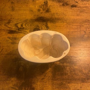 May include: A light-colored, oval-shaped baking mold with a scalloped edge. The interior has a smooth, slightly glossy finish. The mold is sitting on a wooden surface with a visible wood grain.