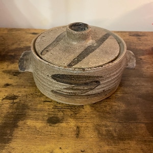 May include: A round, stoneware lidded pot with a textured, speckled finish. The lid has a small handle and dark, diagonal brushstrokes. The pot has two side handles and similar dark brushstrokes around the base. The pot is on a wooden surface.
