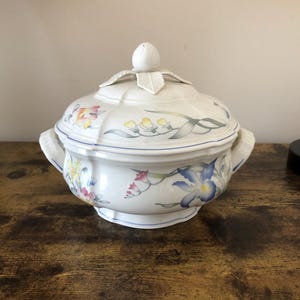 Puede incluir: Una sopera de porcelana blanca con un diseño floral. La sopera tiene una tapa con un pomo en forma de capullo de flor. El diseño floral presenta flores azules, amarillas, rosas y verdes.