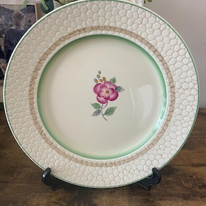 May include: A cream-colored ceramic plate with a floral design. The plate features a pink flower in the center, a green border, and a textured, honeycomb-like rim. The plate is displayed on a black stand.