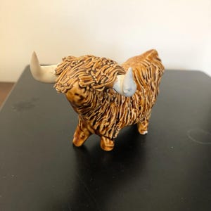 Może przedstawiać: Ceramiczna figurka brązowej krowy szkockiej górskiej z białymi rogami. Krowa stoi na czterech nogach i ma szorstkie futro.