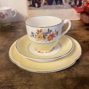 Puede incluir: Juego de taza y platillo de té vintage. La taza tiene un interior blanco, borde negro y base amarilla, adornada con un estampado floral. El platillo es amarillo con borde negro. Una segunda taza es visible.