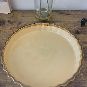 Op de afbeelding: Een ronde, lichtgele keramische taartvorm met een geschulpte rand en een bruine rand. De schaal staat op een houten oppervlak, met een heldere glazen vaas op de achtergrond.