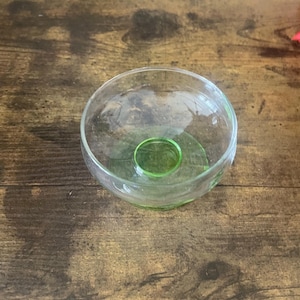 May include: A clear glass bowl with a rounded shape and a green base. The bowl has a small, circular indentation in the centre of the green base. The glass is transparent, allowing light to pass through.