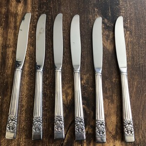 Puede incluir: Seis cuchillos de cena de plata con mangos ornamentados. Los cuchillos tienen un acabado pulido y están dispuestos en una fila sobre una superficie de madera.