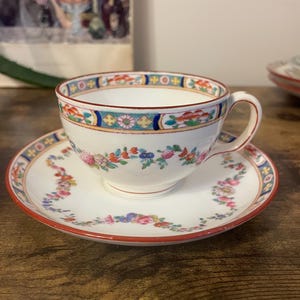 May include: A white teacup and saucer set with a floral design. The cup and saucer have a red trim and a colourful floral pattern. The teacup has a handle and is sitting on the saucer.
