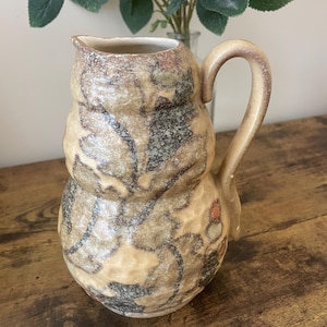 Könnte beinhalten: Ein beigefarbener Keramikkrug mit strukturierter Oberfläche und gebogenem Griff. Der Krug weist ein Muster aus dunkelgrauen und roten floralen Designs auf. Der Krug ist ein dekorativer Gegenstand.