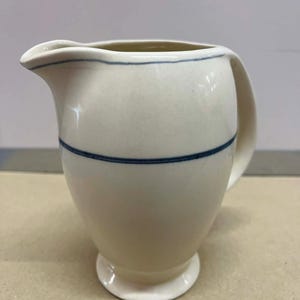 Puede incluir: Jarra de cerámica color crema con una franja azul en el centro y otra en el borde. La jarra tiene un asa y un pico para verter. La base es un pequeño pedestal redondeado.