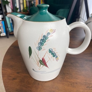 Puede incluir: Tetera de cerámica blanca con tapa y asa verdes. La tetera está decorada con un diseño floral en tonos azules, verdes y rojos. La tetera está sobre una mesa de madera.