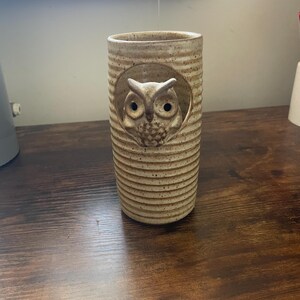 May include: A cylindrical ceramic vessel with a speckled beige finish and horizontal ribbed texture. A sculpted owl figure is integrated into a circular cutout near the top of the piece, featuring detailed facial features and dark, hollow eyes.