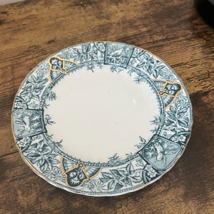 May include: A white and teal decorative plate with a gold rim. The plate features a detailed floral and geometric pattern. The plate is round and appears to be made of ceramic.
