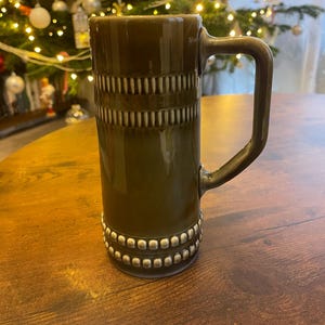 May include: An olive-green ceramic mug with a large handle. The mug has horizontal bands of textured, vertical lines and a beaded design at the base. The mug is on a wooden table. The mug is approximately 15 cm tall.