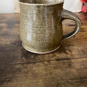 May include: A handcrafted ceramic mug with a speckled, earthy green glaze. The mug features a rounded body, a robust handle, and a slightly flared rim. The mug is placed on a dark wooden surface.