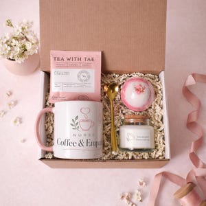 Może przedstawiać: Pudełko prezentowe zawierające biały kubek z różowym uchem i napisem Nurse Coffee & Empathy, różową saszetkę herbaty Tea With Tae, złotą łyżeczkę, okrągłą różową kulę do kąpieli oraz małą świecę w szklanym słoiczku z korkową pokrywką i etykietą.