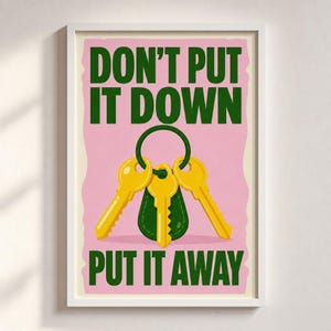 May include: A rectangular wall art print featuring a pink background with dark green text that reads DON'T PUT IT DOWN PUT IT AWAY. In the centre, there is a dark green keychain fob attached to three yellow metal keys on a circular ring.