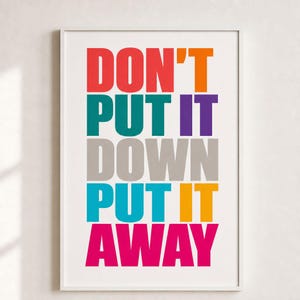 Op de afbeelding: Een rechthoekige wandprint in een witte lijst met de tekst DON'T PUT IT DOWN PUT IT AWAY in dikke, schreefloze hoofdletters. De woorden worden weergegeven in rijen met verschillende kleuren, waaronder rood, oranje, blauwgroen, paars, grijs, blauw en roze.