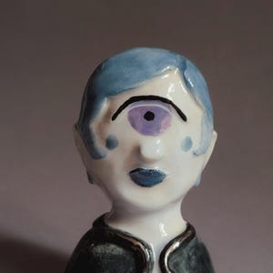 Könnte beinhalten: Eine Keramikfigur, die einen Zyklopen mit hellblauem Haar, einem einzelnen violetten Auge und blauen Lippen darstellt. Die Figur hat ein weißes Gesicht mit kleinen blauen Wangenpunkten und trägt ein dunkles, strukturiertes Kleidungsstück mit silberfarbenem Rand.