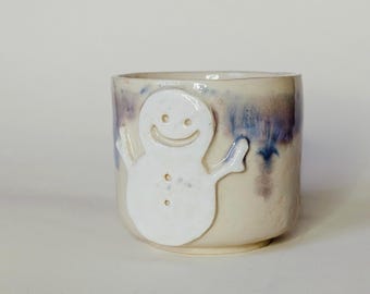 May include: A ceramic mug with a glossy off-white finish and a raised, white snowman figure on the front. The upper rim features a blended wash of blue and purple glaze that drips down the sides, creating a textured, hand-crafted appearance.