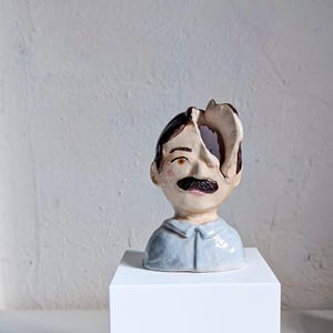 May include: A ceramic sculpture of a man's head with a mustache and a portion of the head missing. The sculpture is painted with brown, black, pink, and blue tones. The head sits on a light blue shirt and is displayed on a white pedestal.