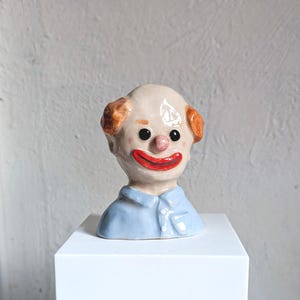 May include: A ceramic clown bust with a pale complexion, red lips, and orange hair. The clown has black eyes and a pink nose. It is wearing a light blue collared shirt. The bust is displayed on a white, square pedestal.