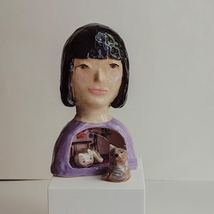 May include: A ceramic bust of a person with black hair and a purple shirt. The torso features a hollow cutout revealing a miniature interior scene with a small cat figurine resting inside. A separate small brown dog figurine sits in front of the bust.