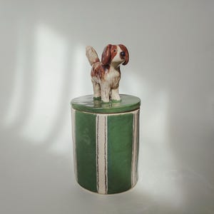 May include: A cylindrical ceramic canister with a green and white vertical striped pattern. The lid features a sculpted, hand-painted brown and white dog figurine standing on top. The canister has a glossy finish and a smooth, rounded shape.