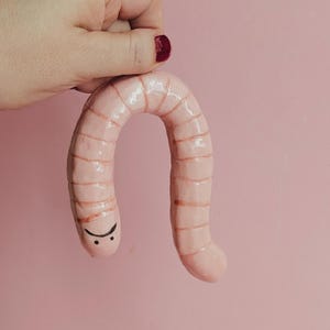 May include: A light pink ceramic worm figurine with a glossy finish and segmented body. The worm features a hand-painted black face with two small dots for eyes and a curved line for a mouth. The object is curved in an inverted U-shape.