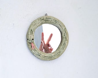 May include: A round wall mirror framed by a sculpted, pale green ceramic snake with a textured surface, dark spots, and a protruding pink tongue. The snake's body forms the circular border of the mirror, which reflects a hand making a peace sign.
