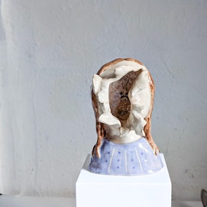 May include: A ceramic sculpture of a head with a missing face. The head has brown hair and a blue dress with white polka dots. The sculpture is displayed on a white pedestal.