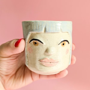 Könnte beinhalten: Eine Keramik-Tasse mit Gesichtsdesign. Die Tasse hat ein hellblaues, haarähnliches Design, schwarze Augenbrauen und rosa Wangen. Das Gesicht hat einen neutralen Ausdruck mit rosa Lippen und einer kleinen Nase. Die Tasse wird vor einem rosa Hintergrund gehalten.