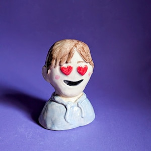 May include: A ceramic sculpture of a person's head and shoulders, with brown hair and red heart-shaped eyes. The figure is wearing a light blue hoodie, with pink cheeks and a smiling black mouth. The background is a solid purple.